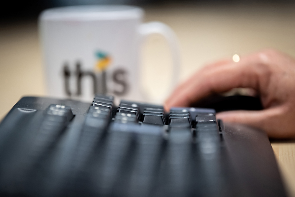 Computer keyboard and mouse with the THIS branded white mug in the background.