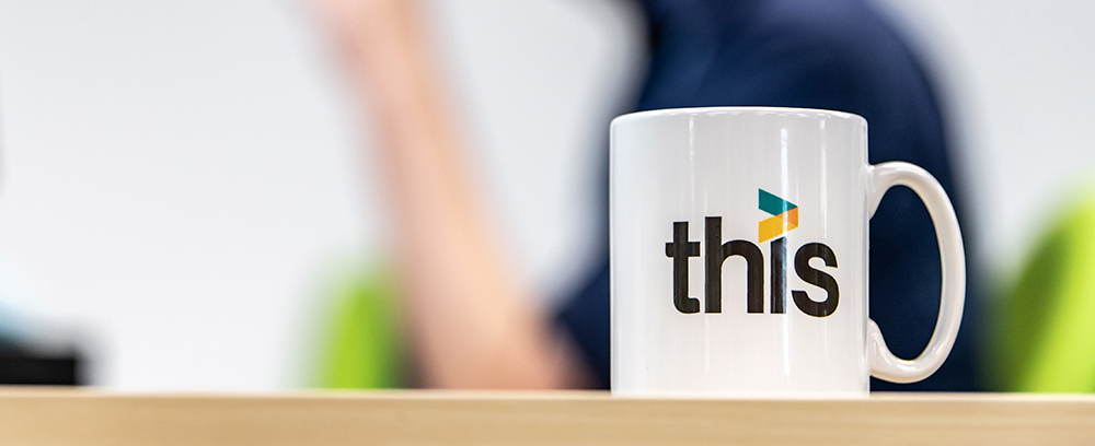 THIS branded white mug on a desk with a man behind working on a computer.