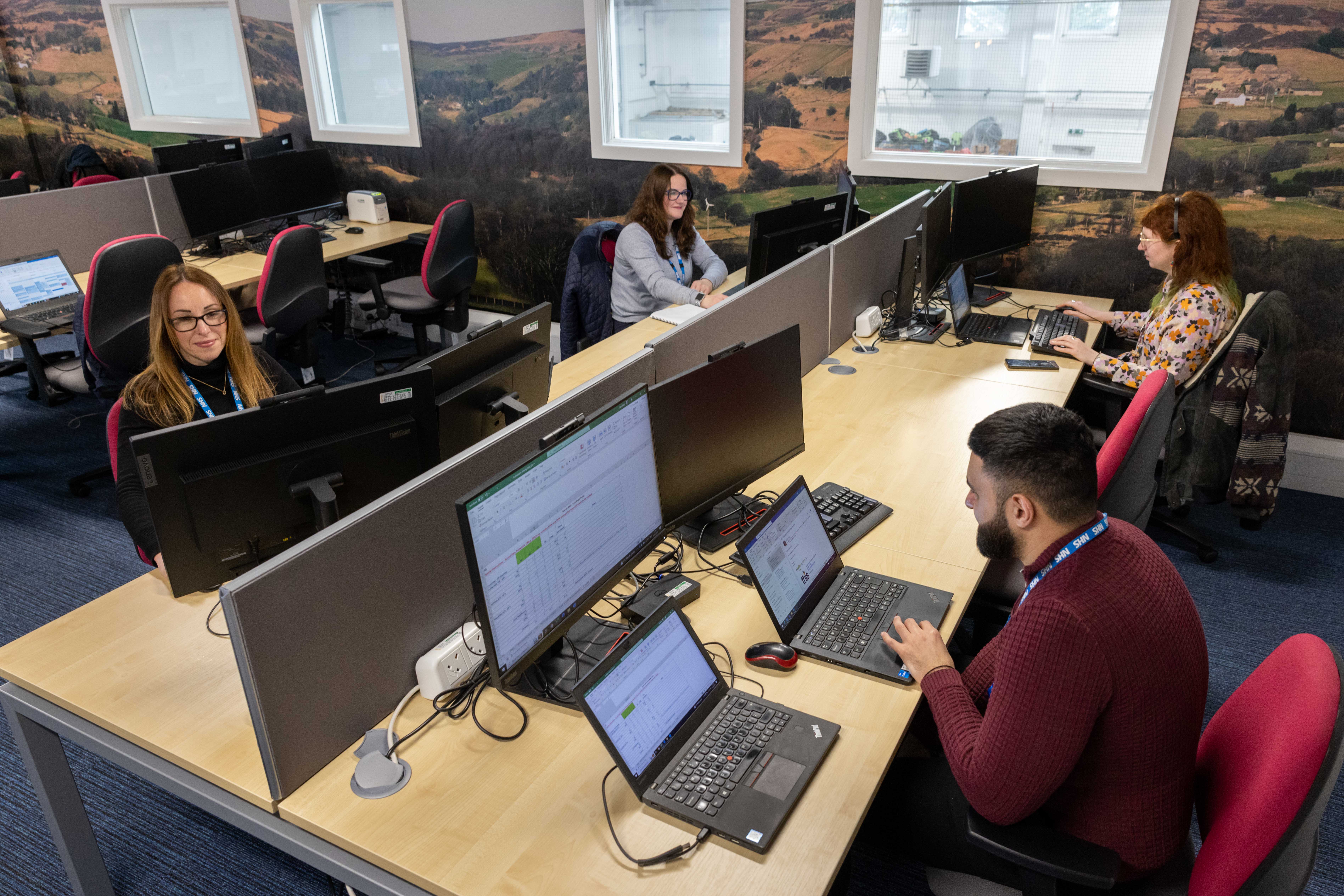 Male and Female colleagues sat in Elland's Hot Desking room working on their laptops connected to external monitors.