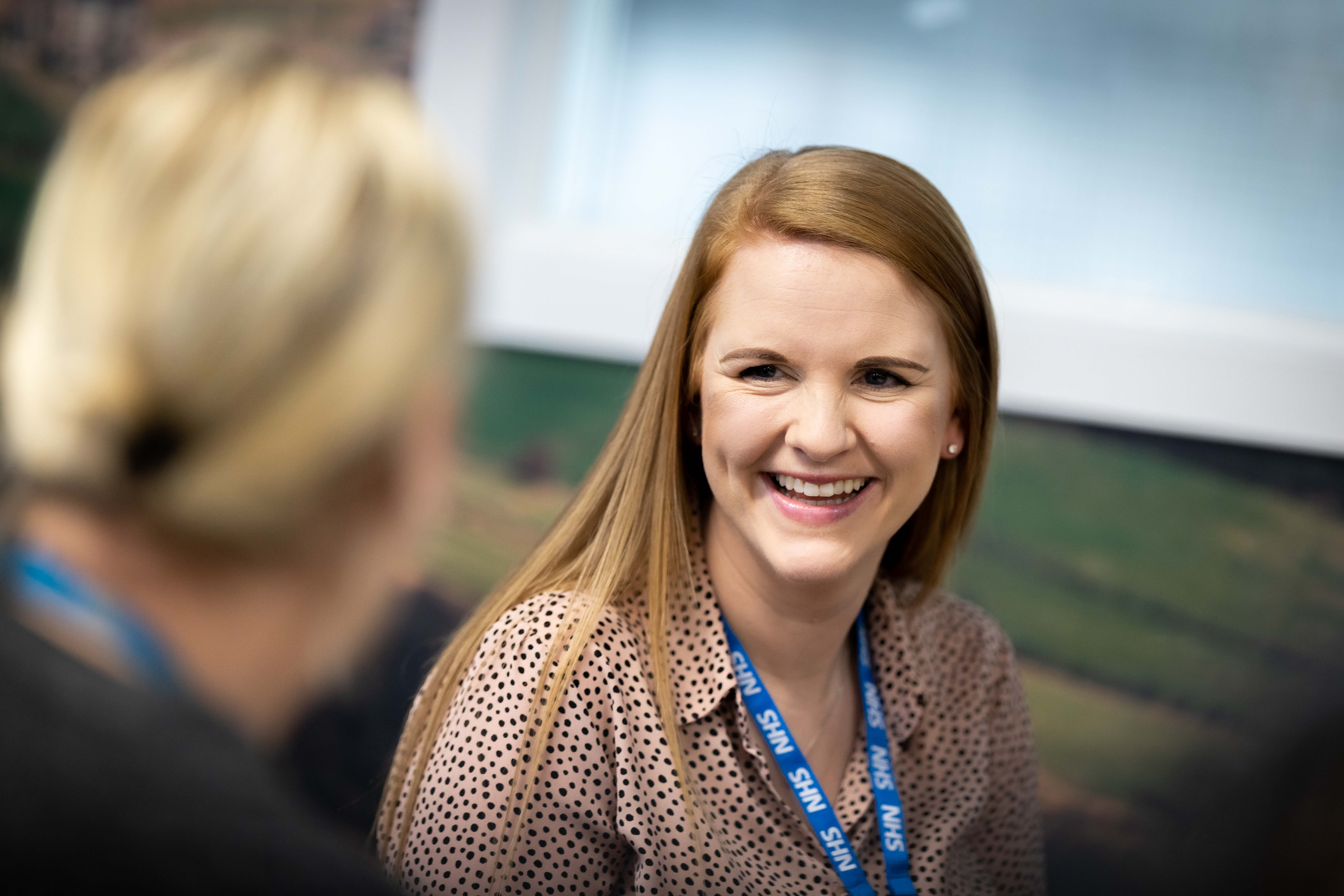 Women wearing a NHS badge smiling in conversation with another women in the Elland office.